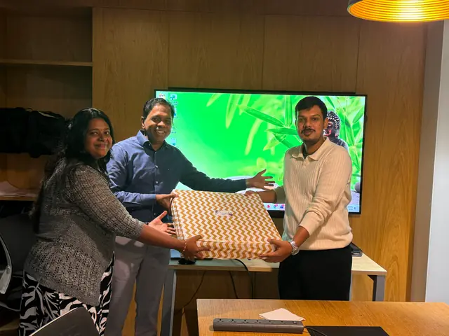 Two colleagues handing a large gift-wrapped box to a newly married teammate in the office, all three smiling