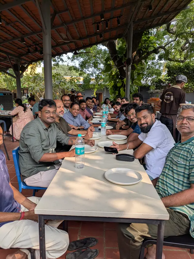 A long outdoor lunch table under a tiled roof and a big tree, the team smiling at the camera